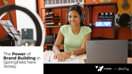 Podcast Brand Building: Female podcaster recording indoors with a computer, papers, and pen on the table, smiling at the camera.