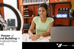 Podcast Brand Building: Female podcaster recording indoors with a computer, papers, and pen on the table, smiling at the camera.