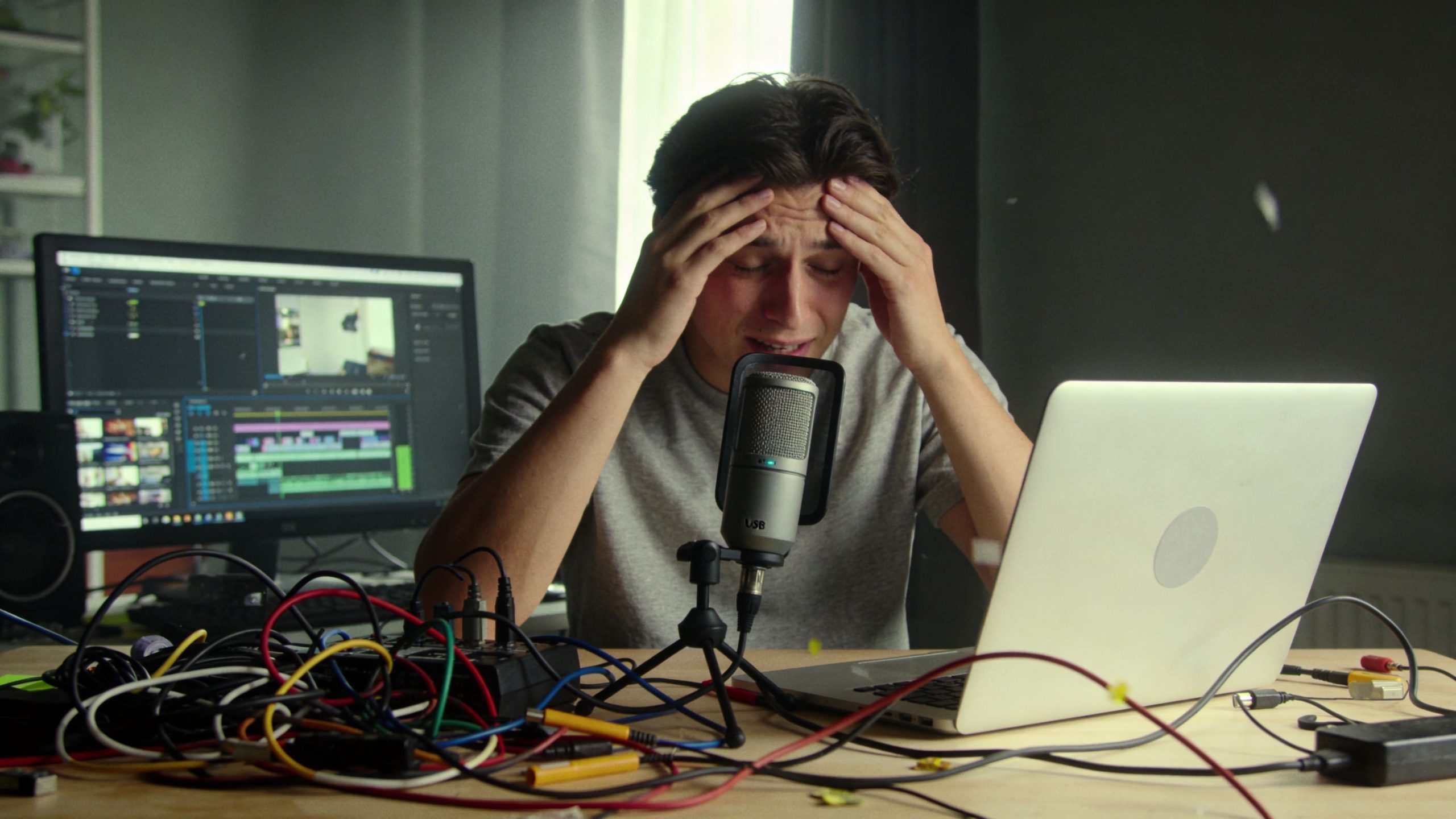 A frustrated content creator holding his head while working with audio and video editing equipment at home.